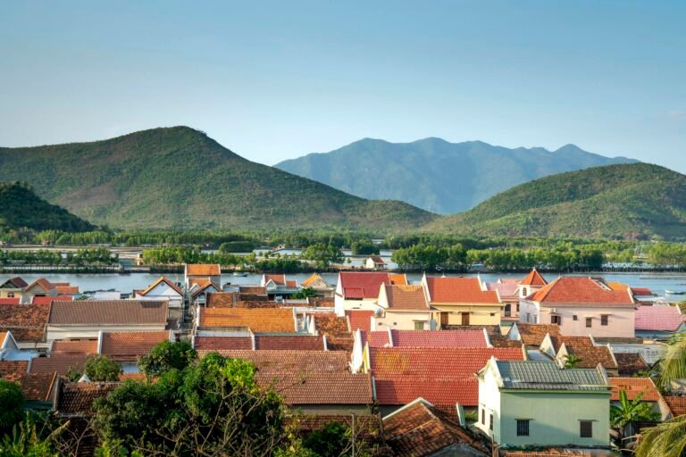 A picturesque village set against lush mountains and a serene river under a clear summer sky.