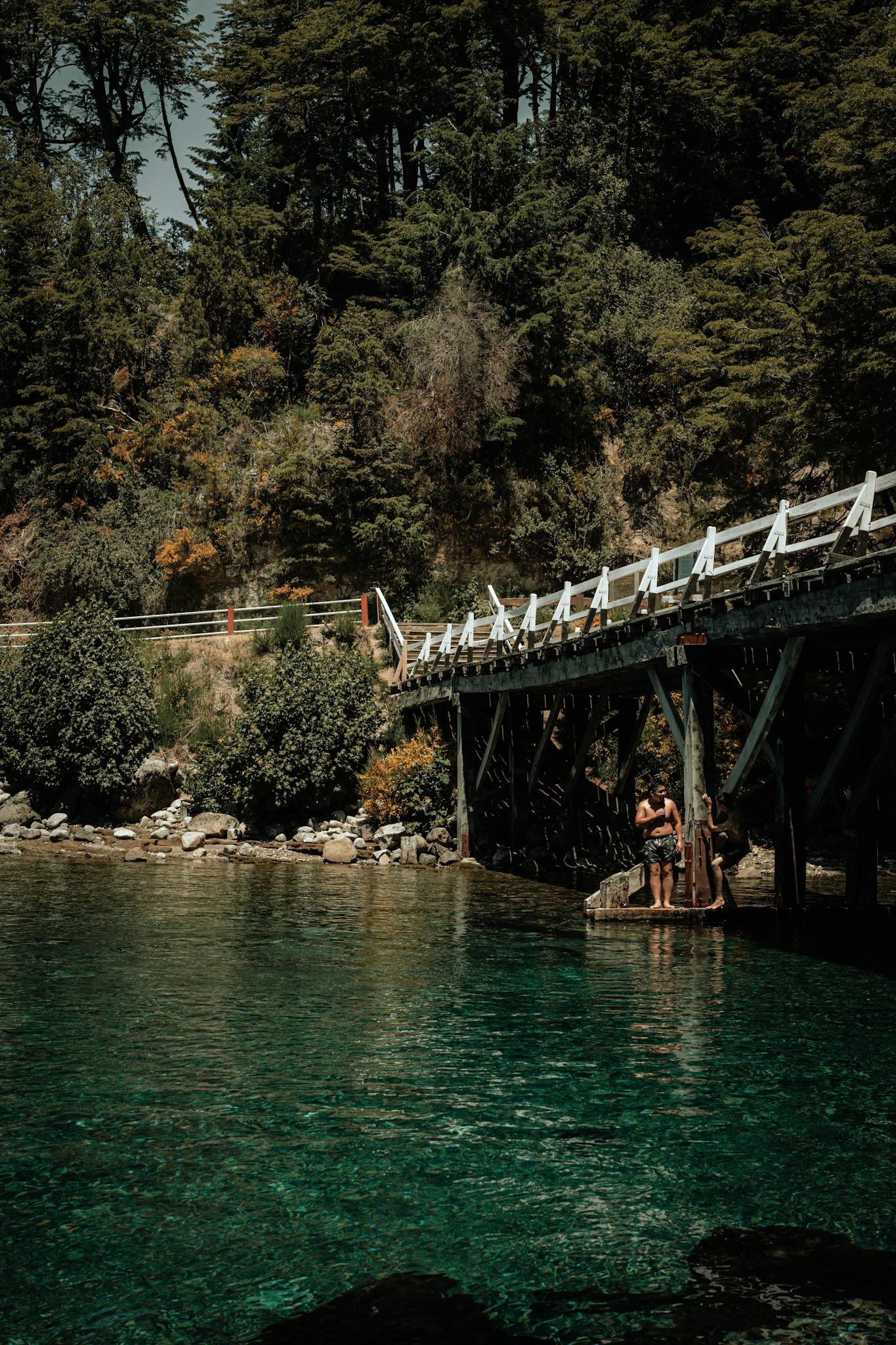 A tranquil scene of people relaxing by a river under a wooden bridge in Villa La Angostura, Argentina.