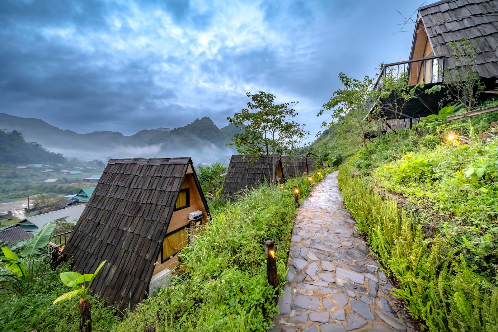 Scenic view of A-frame cabins along a stone path in a lush mountainous area with misty skies.