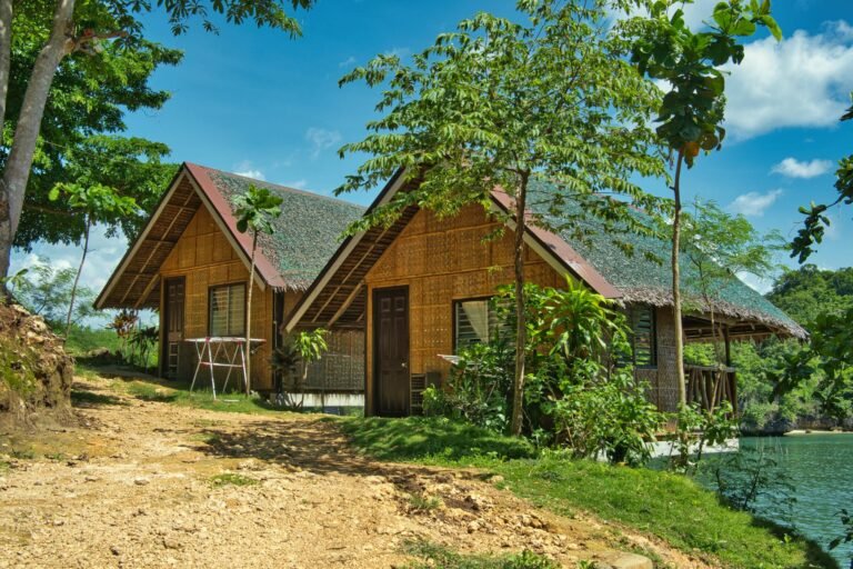 Serene bamboo cottages by the lake, surrounded by lush green trees and a clear blue sky.